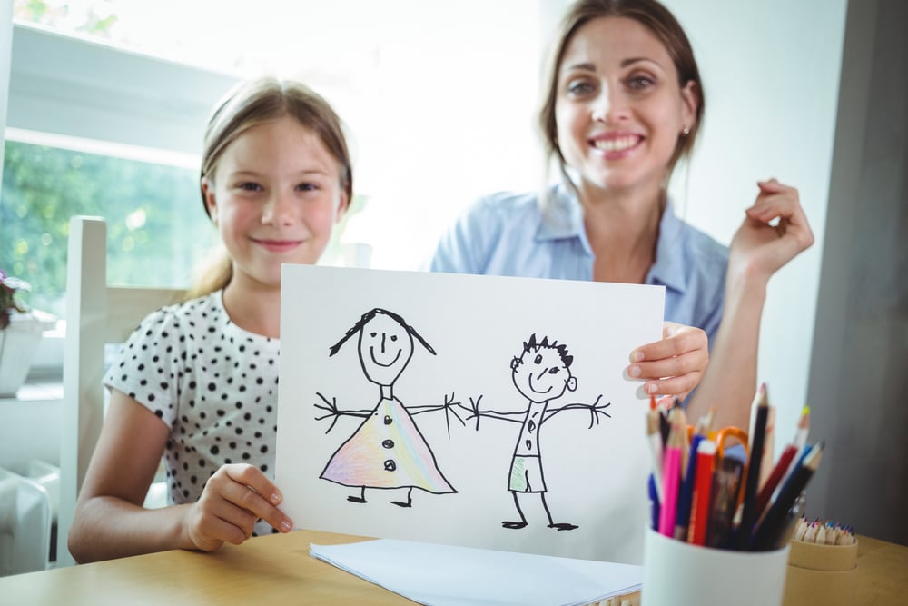 Portrait of mother and daughter sitting at table and daughter showing her drawing